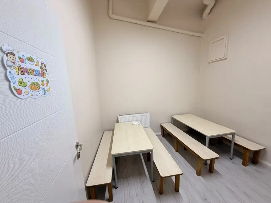 Kapısında Yemekhane yazısı bulunan, ahşap masa ve banklarla düzenlenmiş okul yemekhanesi